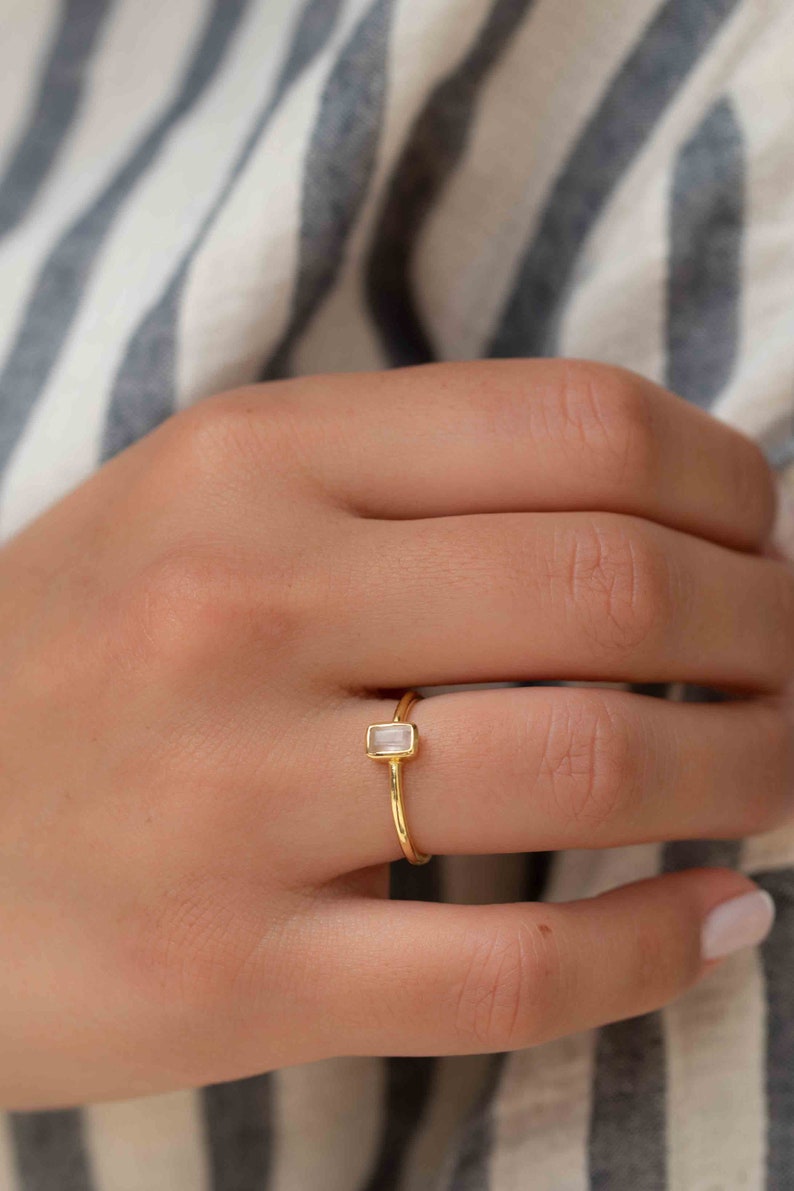 May include: A gold ring with a rectangular clear gemstone. The ring is on a finger.