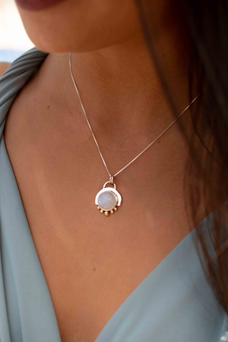 May include: A silver chain necklace with a moonstone pendant. The pendant is surrounded by a silver frame and has small gold beads around the bottom.