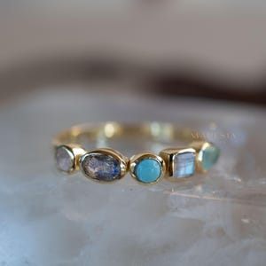May include: Gold ring with a row of five oval and square shaped gemstones. The gemstones are a mix of blue, grey, and white.
