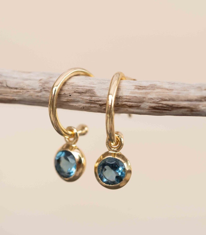 May include: A pair of gold hoop earrings with a small blue gemstone dangling from each hoop.