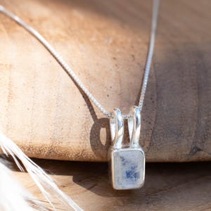 Puede incluir: Un collar de cadena de plata con un colgante rectangular de piedra lunar. El colgante está engastado en un marco de plata y tiene un delicado brillo iridiscente.
