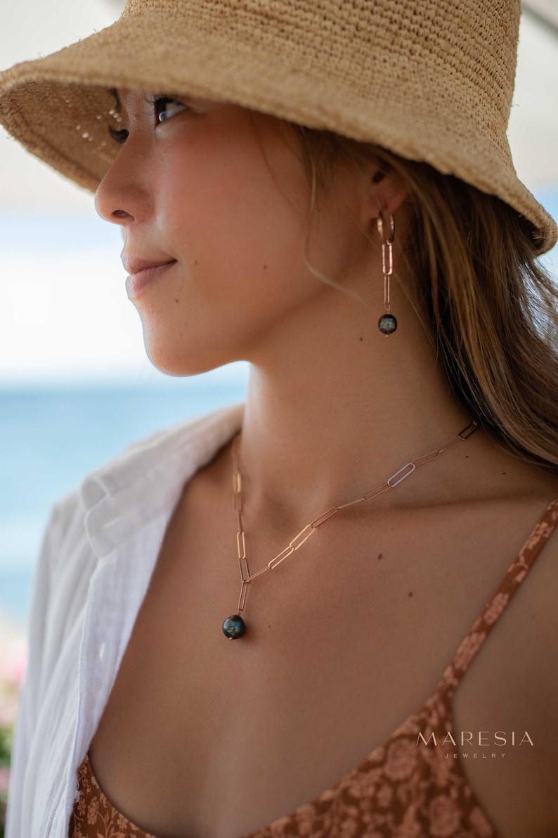 K&ouml;nnte beinhalten: Eine Frau tr&auml;gt einen Strohhut und ein wei&szlig;es Hemd mit einer goldenen Kettenkette und Ohrringen mit einem schwarzen Perlenanh&auml;nger. Die Kette und die Ohrringe sind von der Marke "Mares&iacute;a Jewelry".