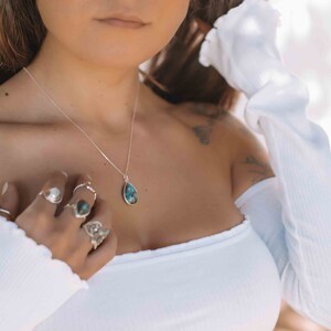 May include: A silver necklace with a teardrop-shaped blue gemstone pendant. The necklace is worn by a person with multiple silver rings on their fingers.