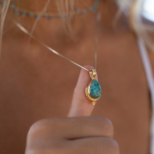 May include: Gold-toned pendant necklace featuring a teardrop-shaped turquoise stone. The pendant is suspended from a delicate chain. A matching turquoise bead choker is visible in the background. The necklace is held up by a person's thumb.