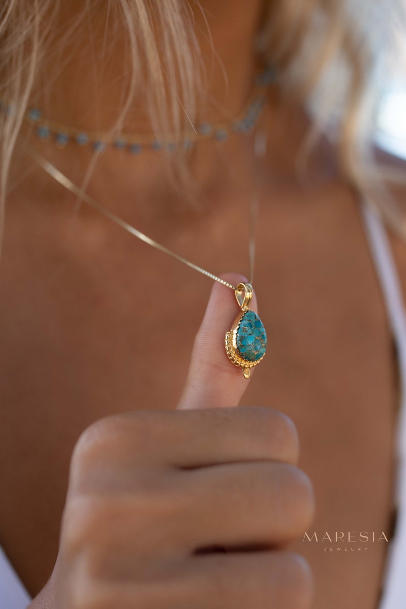 May include: Gold-toned pendant necklace featuring a teardrop-shaped turquoise stone. The pendant is suspended from a delicate chain. A matching turquoise bead choker is visible in the background. The necklace is held up by a person's thumb.