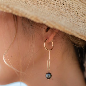 K&ouml;nnte beinhalten: Goldene Creolen mit einer zarten Kette und einer schwarzen Perle, die am Ende baumelt.  MARESIA JEWELRY ist in wei&szlig;er Schrift auf braunem Hintergrund gedruckt.
