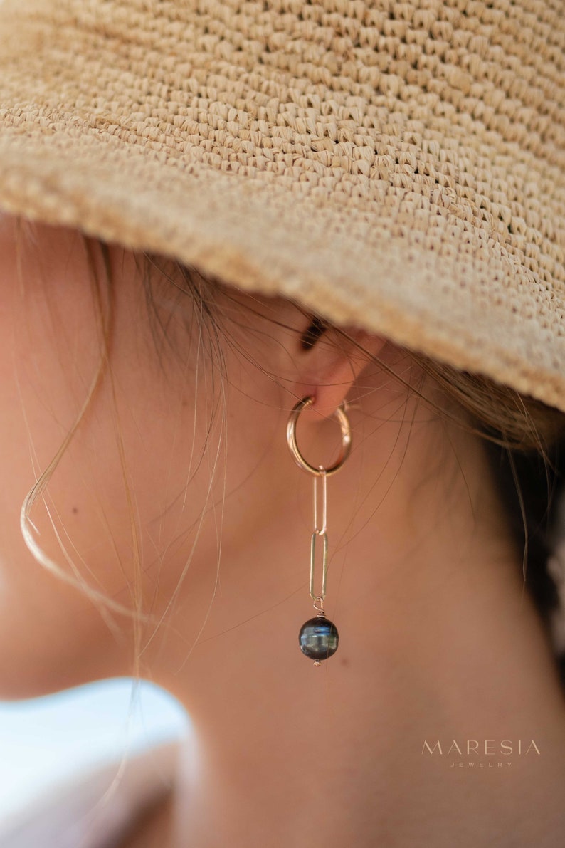 K&ouml;nnte beinhalten: Goldene Creolen mit einer zarten Kette und einer schwarzen Perle, die am Ende baumelt.  MARESIA JEWELRY ist in wei&szlig;er Schrift auf braunem Hintergrund gedruckt.