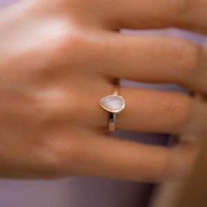 May include: A gold ring with a teardrop-shaped moonstone gemstone. The ring is on a person's finger.