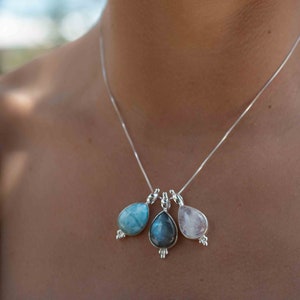 May include: A silver chain necklace with three teardrop-shaped pendants. The pendants are made of blue, gray, and pink stones.