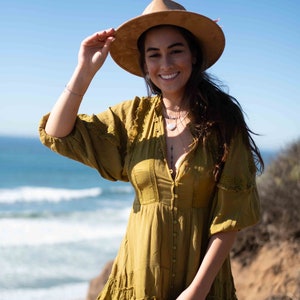 May include: A woman wearing a mustard-colored dress and a wide-brimmed hat. The dress has a button-down front and ruffled hem. The background features the ocean and a clear blue sky.