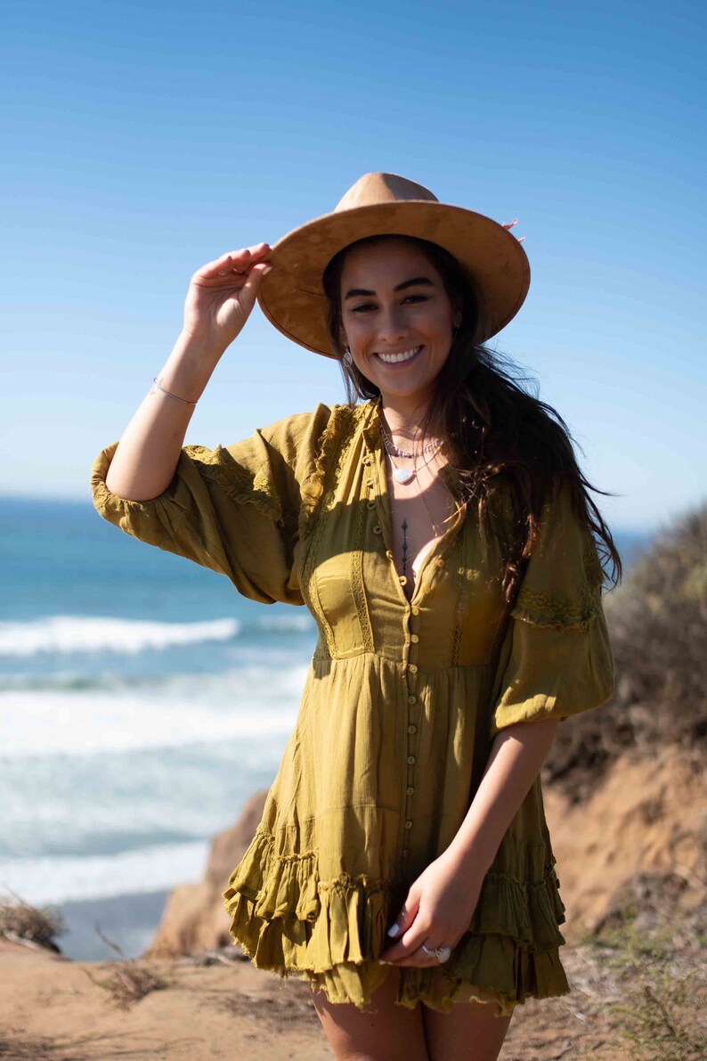 May include: A woman wearing a mustard-colored dress and a wide-brimmed hat. The dress has a button-down front and ruffled hem. The background features the ocean and a clear blue sky.