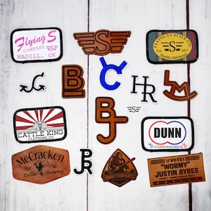 Peut inclure: Un assortiment d'écussons en cuir et en tissu avec divers motifs et textes. Les écussons présentent des logos, des noms de marques et des textes tels que "Flying S Company", "Cattle King Clothing Co." et "Squatchsprayz". Les couleurs incluent le marron, le noir, le blanc, le rouge et le bleu.