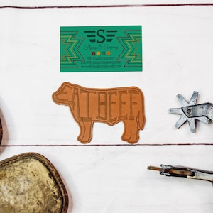 May include: A light brown leather cow-shaped cutout with the words "EAT BEEF" cut out of the body. A green business card with gold text and a logo is above the cow. A metal spur and tool are to the right.