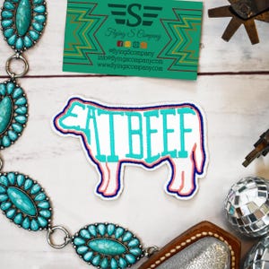 May include: A white patch in the shape of a cow with the words "EAT BEEF" in turquoise. The patch has a pink and blue outline. A turquoise and silver belt and a business card are also visible.