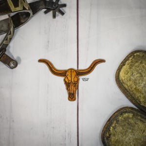 May include: A wooden sticker of a longhorn skull with the initials "S=" on a white wooden surface. A spur and a leather boot sole are also visible.