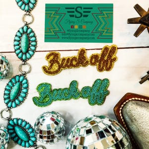 May include: Two iron-on patches with the text "Buck off" in a cursive font. The top patch is gold with brown stitching, and the bottom patch is green with turquoise stitching.
