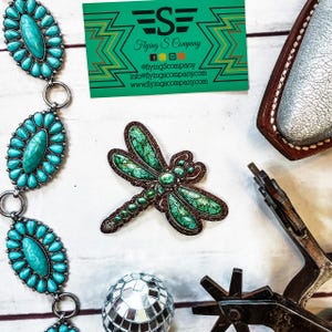 May include: An overhead shot features a turquoise and silver belt, a green and brown dragonfly-shaped accessory, a disco ball, and a business card. A spur and a leather item are also visible. The business card has the text "Flying S Company".