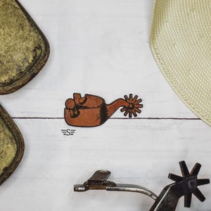 May include: A brown leather spur with a star-shaped rowel and a decorative design. The spur is a western-themed accessory, often used in equestrian activities. The image also includes a logo with the letters "ES".