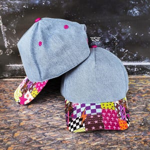 May include: Two denim baseball caps with pink button details. The brims feature a colorful patchwork design with geometric shapes, stars, and lightning bolts. The caps are casual and stylish, suitable for everyday wear.