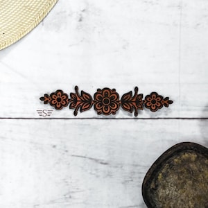 May include: A flat, horizontal decorative cutout featuring a symmetrical floral and leaf pattern in black and burnt orange. The design consists of a central flower flanked by two sets of leaves and smaller flowers on each end, with a small logo.