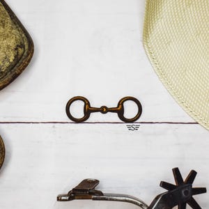 May include: A bronze-colored horse bit with two circular rings connected by a central bar. The bit is centered on a white wooden surface, with other equestrian-themed items partially visible.