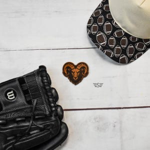 May include: A black leather baseball glove with white stitching, a brown leather patch featuring a ram head design, and a baseball cap with a black and white pattern of footballs. The cap has a cream-colored front panel and a black brim.