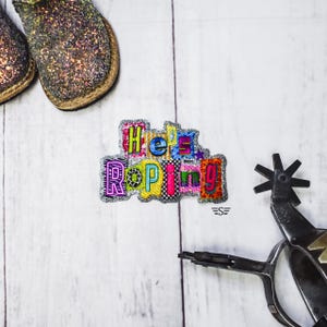 May include: An embroidered patch featuring the text "He's Roping" in colorful, patterned block letters with a silver glitter border. The patch is positioned between a pair of dark, glittery boots and a metal western spur with a rowel.