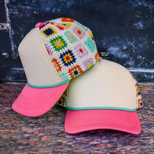 May include: Two trucker hats featuring a cream-colored front panel, a vibrant multi-colored crochet granny square pattern on the back panels, a bright pink brim, and a thin teal cord trim separating the front panel from the brim.