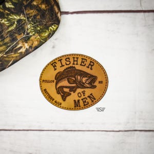 May include: A brown leather patch with the words "FISHER OF MEN" and a fish illustration. The text "FOLLOW ME" and "MATTHEW 4:19" are also included. A camouflage pattern is visible in the upper left corner.
