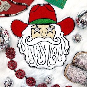 May include: A white felt Christmas ornament with a red and green Santa Claus wearing a cowboy hat and sunglasses. The ornament has a glittery finish and is surrounded by silver disco balls and red beaded jewelry.