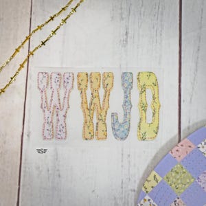 May include: A fabric patch with the letters "WWJD" in a decorative font. The letters are filled with floral patterns in pastel colors, including yellow, pink, and blue. A gold chain and a patchwork design are also visible.