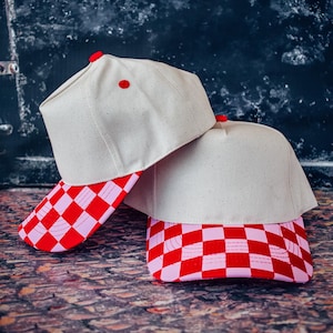 May include: Two beige baseball caps with red and pink checkered brims. Each cap has a red button on top. The caps are stacked on top of each other, showcasing the design and color scheme.