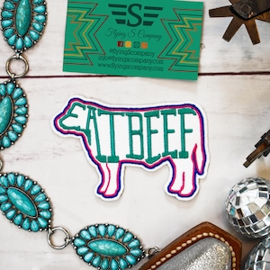 May include: White embroidered patch with a cow silhouette and the text "EAT BEEF" in green lettering. The patch has pink and blue outlines.