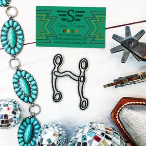 May include: A silver and black embroidered patch of a horse bit, with the text "Flying S Company" on a green and yellow business card in the background.