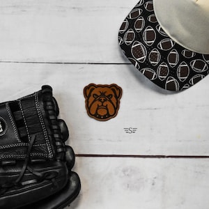 May include: A brown leather patch featuring a bulldog head design, positioned between a black baseball glove and a baseball-themed cap with a black and white pattern. The small logo text ES is visible near the center of the composition.