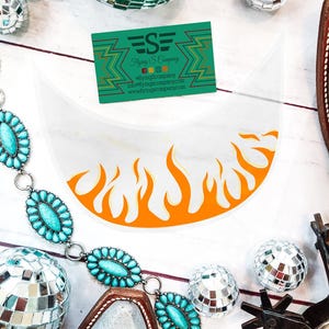 May include: A white curved visor featuring an orange flame graphic along the bottom edge. A green business card for Flying S Company is positioned above the visor. A turquoise stone chain, silver disco balls, and a metal spur are also visible.