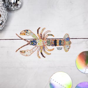 May include: A colorful lobster sticker with a patchwork design. The lobster's body is made up of various floral and patterned fabrics in shades of blue, yellow, and green. The sticker has a white background and the initials "S E" are visible.