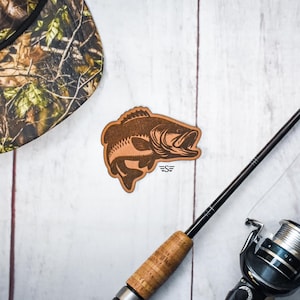 May include: A brown bass fish-shaped wooden cutout with an open mouth, next to a fishing rod and reel. A camouflage hat is in the upper left corner. The fish cutout has the initials "SE" below it.