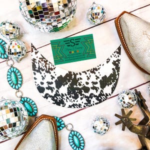 Puede incluir: Una diadema de tela con estampado de piel de vaca blanca y negra con un collar de cuentas turquesa y plata, una bota plateada y una tarjeta de visita verde con el texto "Flying S Company" y la dirección del sitio web.