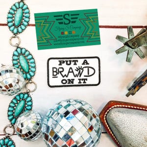 May include: An assortment of western-themed items on a white wooden surface. Includes a turquoise and silver belt, a disco ball, a business card, a patch that says "PUT A BRAND ON IT", and a spur.