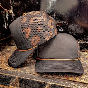 May include: Two black trucker hats. One hat features a leopard print mesh top and a black front panel. The other hat is solid black. Both hats have a curved brim and a gold-colored rope detail along the front.