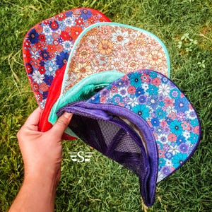 May include: Three trucker hats stacked on top of each other. The top hat is teal with a floral pattern, the middle hat is red with a floral pattern, and the bottom hat is purple with a floral pattern.