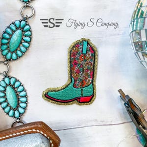 May include: A turquoise and floral cowboy boot patch with gold glitter trim. The boot has a red sole and a turquoise pull tab. The patch is next to a turquoise stone belt and the text "Flying S Company".