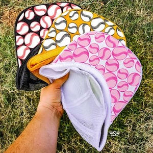 Könnte beinhalten: Eine Sammlung von Baseball-Trucker-Caps. Die Caps haben einen Mesh-Rücken und einen gebogenen Schirm. Die Schirme zeigen Baseballmuster in Schwarz, Gelb und Rosa mit weißen und roten Baseballnähten. Die Caps werden in einer Hand gehalten.