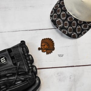 May include: A black leather baseball glove, a baseball cap with a white crown and a black brim featuring a repeating football pattern, and a brown leather patch depicting a stylized figure wearing a headdress and holding a baseball bat.