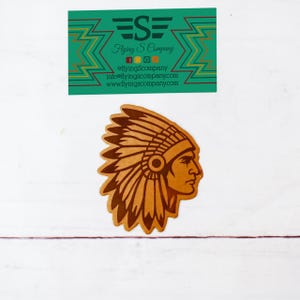 May include: A tan-colored leather patch featuring a profile of a Native American chief wearing a feathered headdress. Above the patch is a green business card with the text "Flying S Company".