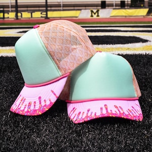 May include: Two trucker hats with mint green front panels and pink brims. The brims feature a pink frosting design with colorful sprinkles. The mesh backings are tan, and the hats have pink piping.