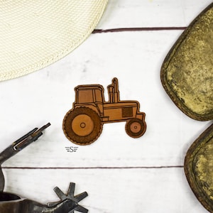 May include: A brown tractor-shaped wooden cutout with a large rear tire and small front tire. The tractor has a detailed design and is set against a white wooden background. The cutout has the letters "ES" below it.
