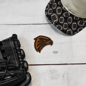 May include: A black leather baseball glove, a brown leather patch featuring an eagle head silhouette, and a black baseball cap with a white football pattern. The patch includes a small logo with the text S flanked by two horizontal lines.