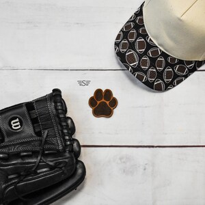 May include: A black leather baseball glove with white stitching, a cream-colored baseball cap featuring a black and white football pattern on the brim, and a brown leather paw print patch with the text S centered between them.
